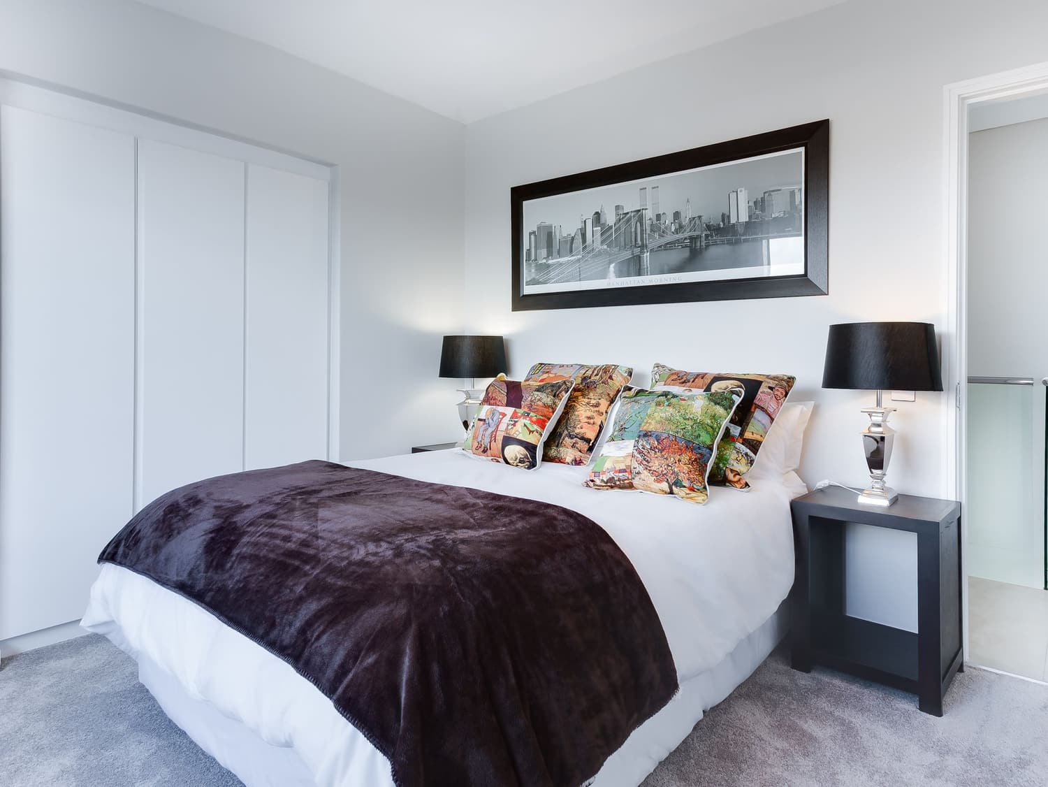 A bedroom with a queen bed, a dark throw and white duvet, four patterned cushions, two black table lamps on dark nightstands, a framed panoramic print, a fitted wardrobe with white sliding doors and fitted gray carpet. Numbered markers identify each item found, listed in the two tables below.