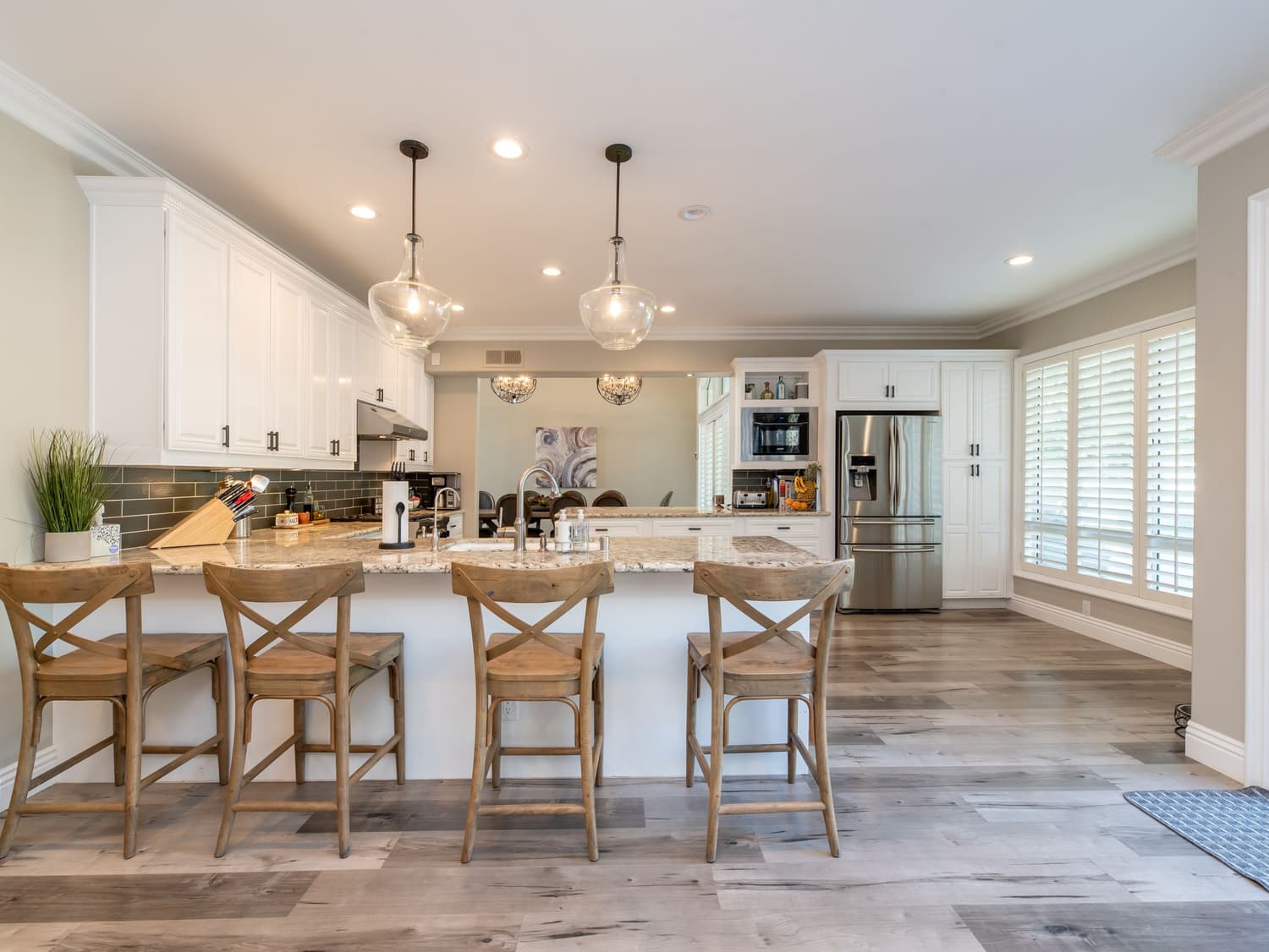 An open-plan kitchen with white shaker cabinetry, a granite island with four wooden bar stools, a stainless French-door refrigerator, a built-in microwave, glass pendant lights, recessed lighting, plantation shutters and wood-look flooring. Numbered markers identify each item found, listed in the two tables below.