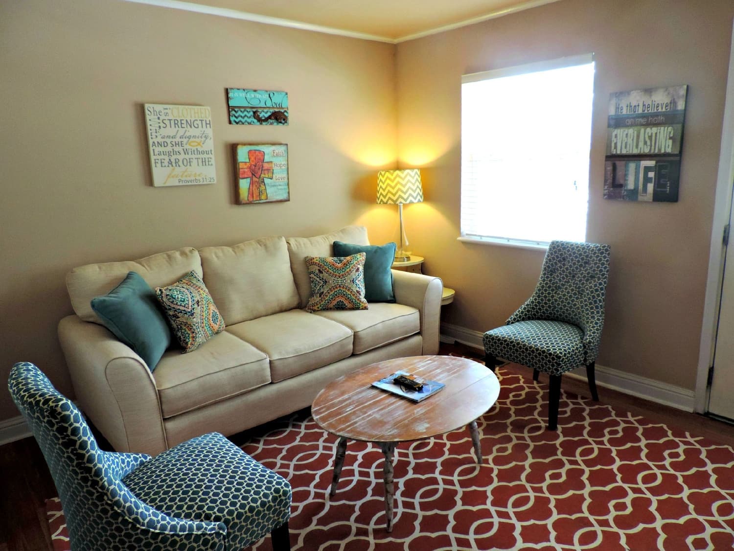 A furnished living room with a beige three-seat sofa, two patterned accent chairs, a round wooden coffee table, a patterned area rug, a table lamp on a side table, framed wall art, a blinded window and hardwood flooring. Numbered markers identify each item found, listed in the two tables below.